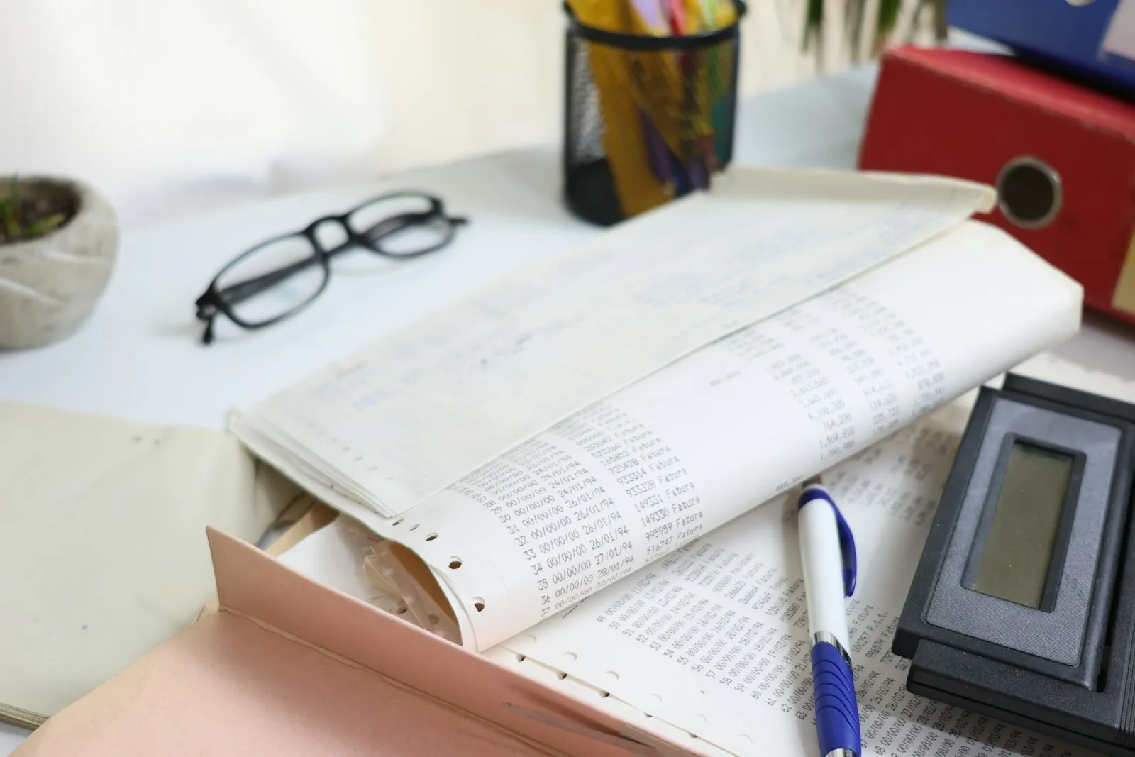A desk with printed ledgers, a pen, reading glasses and a calculator