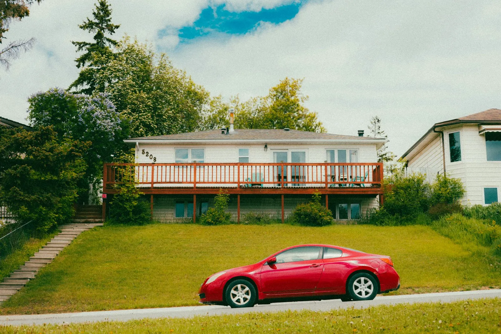 A red coupe parked on a curb in front of a white bungalow with a wooden deck