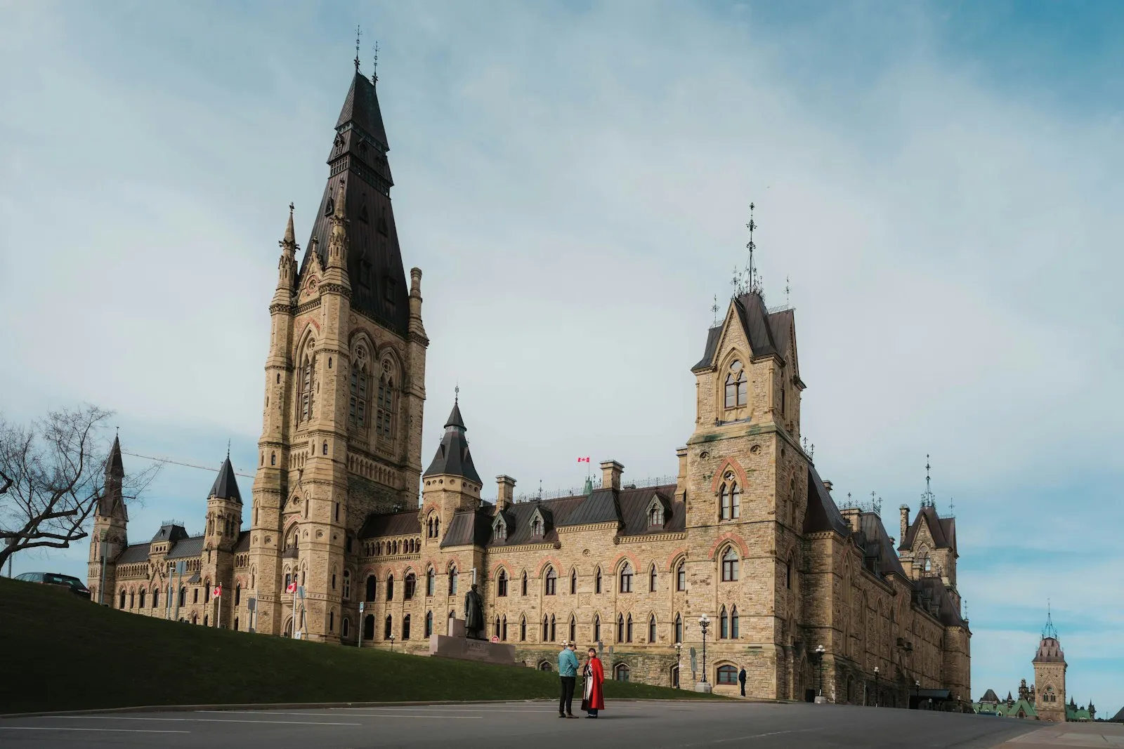 The West Block of Parliament Hill in Ottawa under a blue sky, with two people standing in front