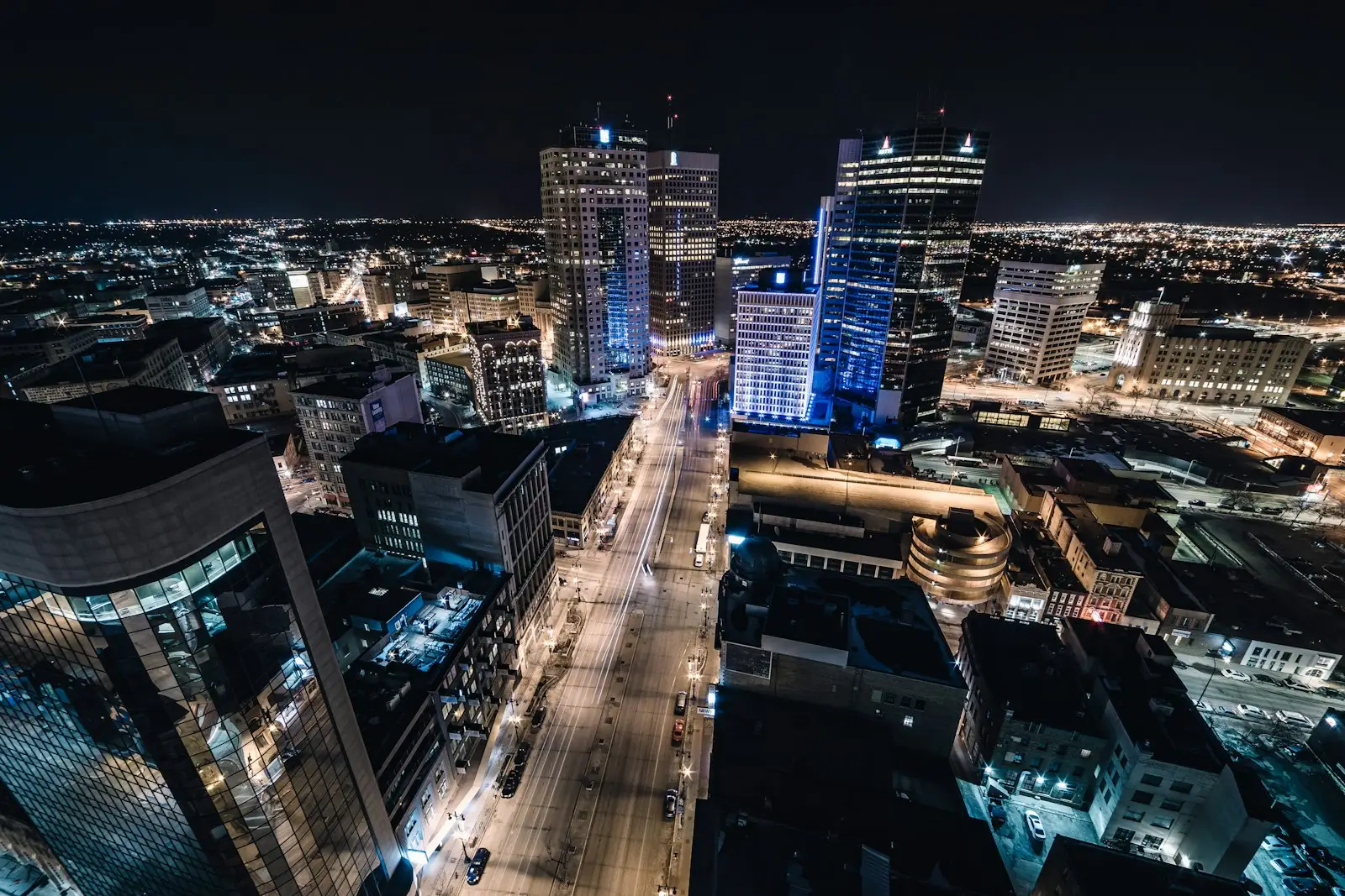 Downtown Winnipeg at night seen from high above, with office towers lit in white and blue and light trails along a wide main street