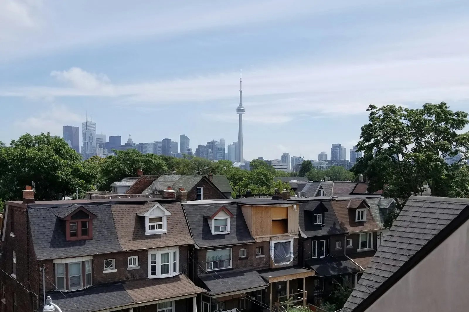 Shingled roofs of older semi-detached houses in a Toronto neighbourhood, with the CN Tower and downtown skyline behind